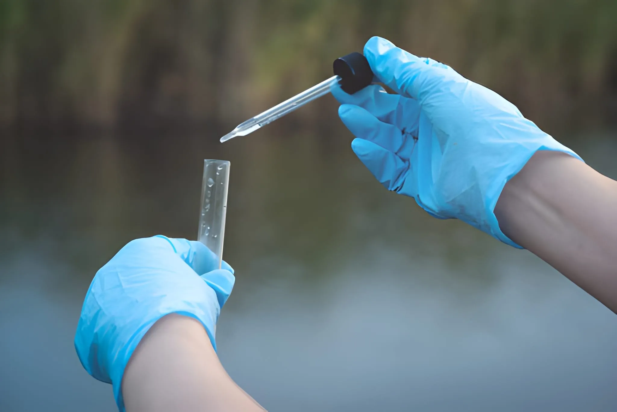 Dua tangan ber-sarung tangan biru sedang melakukan teknik sampling air di lapangan, menggunakan pipet tetes untuk memasukkan sampel cairan ke dalam tabung reaksi dengan latar belakang permukaan air.