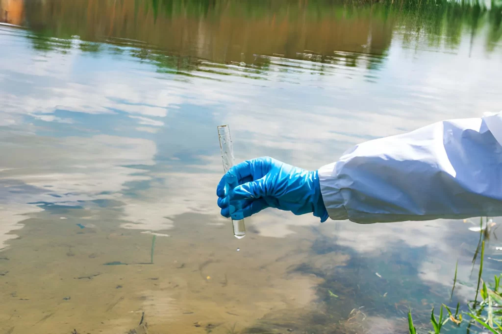 Ilustrasi proses pengambilan sampel air oleh petugas di tepi sungai sebagai contoh penjelasan apa itu pengambil contoh uji air, menggunakan sarung tangan dan tabung sampel untuk analisis kualitas lingkungan