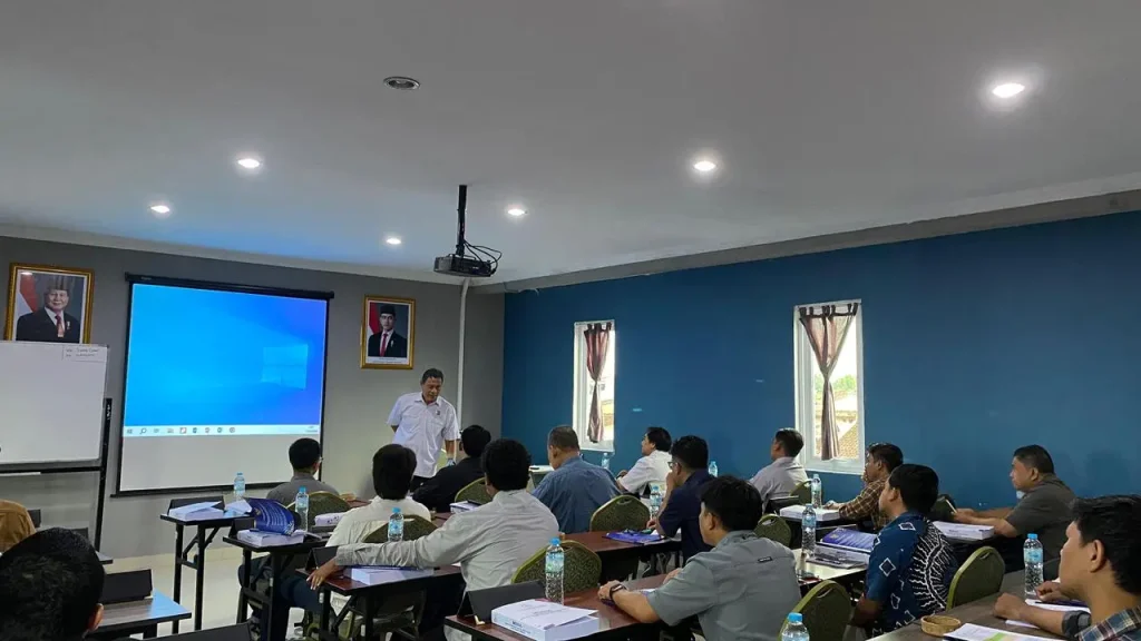 Pembinaan Ahli K3 PUBT Hari Kedua dalam suasana kelas pelatihan, menampilkan instruktur yang sedang memberikan materi di depan layar proyektor kepada peserta yang duduk rapi dengan perlengkapan modul dan botol air, mencerminkan proses pembelajaran yang interaktif dan profesional.