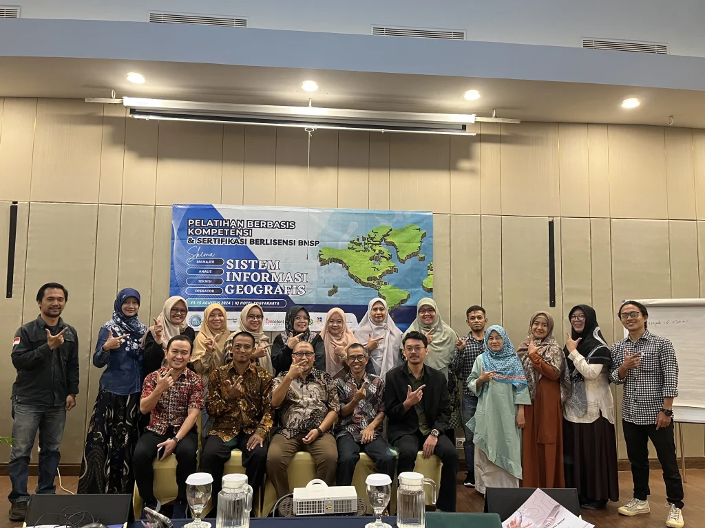 Foto bersama para peserta dan instruktur dalam acara Pelatihan Berbasis Kompetensi & Sertifikasi Berlisensi BNSP untuk Operator Utama GIS di KJ Hotel Yogyakarta. Mereka berpose bersama di depan spanduk acara dengan latar belakang peta dunia